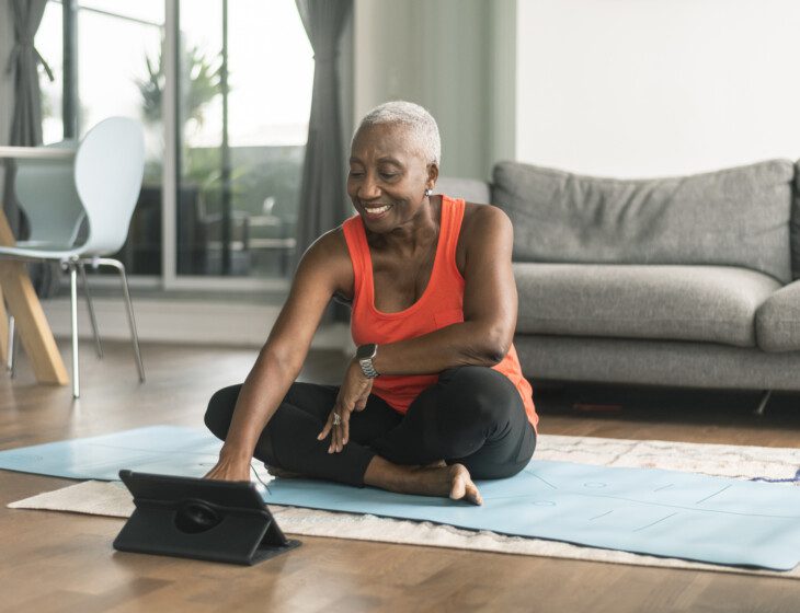 A beautiful senior woman takes an online yoga class. She is interacting with the teacher on her tablet and about to commence her class. She is wearing casual active wear and is taking the class in her lounge room. She is sitting on a blue yoga mat.