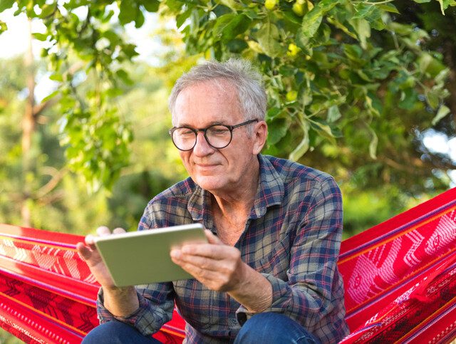 Older adult man sitting on hammock reviewing information on tablet device.