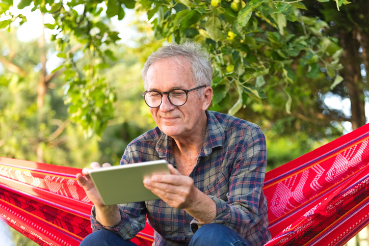 Older adult man sitting on hammock reviewing information on tablet device.